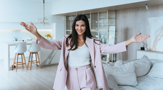 cheerful Asian real estate agent girl in a suit spreads her arms and is invited to inspect the apartment in a new house, looking into the camera and smiling. Realty