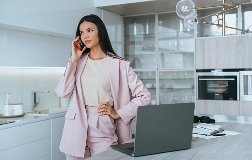 serious business woman, an Asian woman in a pink pantsuit, stands in the kitchen talking on phone next to an open laptop, her day begins, negotiations with partner