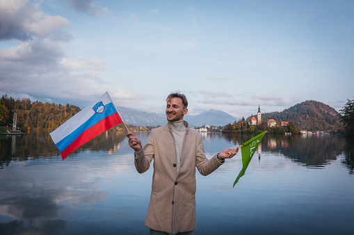 Young male tourist waving Slovenian flags while visiting Lake Bled.