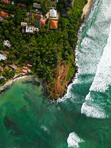 Aerial view from above of lush tropical coastal landscape and vibrant turquoise sea with white-capped waves. Tranquil bay, a traditional village temple, exotic travel destinations nature beauty.