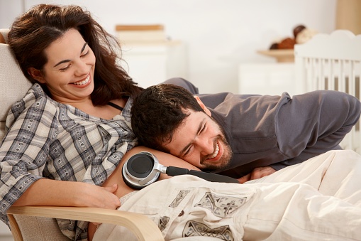 Laughing pregnant woman resting in armchair at home husband holding earphones to naked belly...