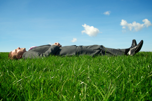 Businessman lays horizontal dreaming in tall green grass under blue sky