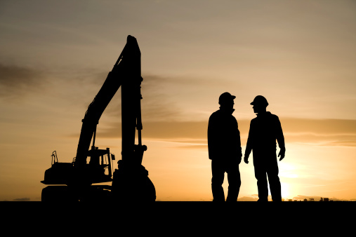 Two silhouetted construction workers and a construction shovel against a sunrise.