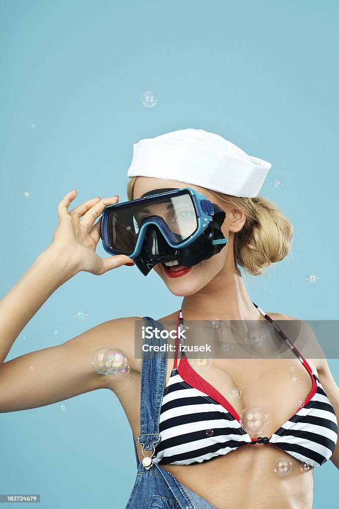 Pin-up style sailor woman looking through scuba mask Young Blond Woman Wearing Striped Bikini and Blue Overalls looking through scuba mask. Standing against blue background. Pin-Up style. Summer portrait. 20-24 Years Stock Photo