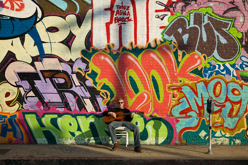 Young man plays classical guitar on sidewalk in front of graffiti brick wall