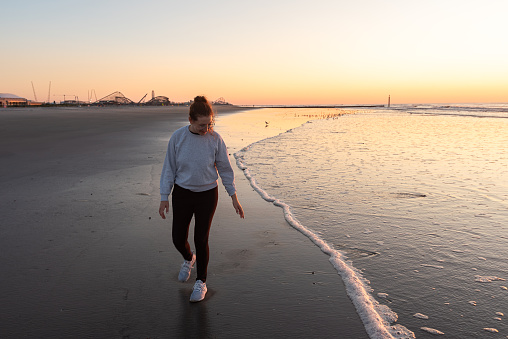 Woman takes a leisurely walk along the sandy shore of Wildwood, New Jersey, USA, as the sun sets over the glistening Atlantic Ocean. Her smile mirrors the warmth of the fading sunlight, creating a delightful and peaceful coastal scene