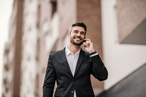A cheerful businessman is walking on the city street and talking on the phone.