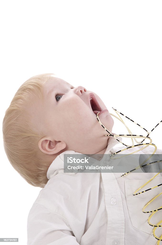Baby boy with mouth open and head back playing with slinky. One-year-old boy looks up with slinky caught in mouth. Vertical studio shot on white. Metal Coil Toy Stock Photo
