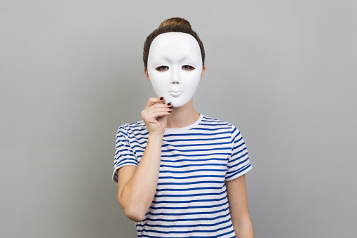 Portrait of unknown anonymous woman wearing striped T-shirt covering her face with white mask, hiding personality, conspiracy and privacy. Indoor studio shot isolated on gray background.