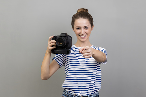 Portrait of joyful woman photographer in T-shirt holding professional digital dslr camera and pointing at you with toothy smile, enjoying her job. Indoor studio shot isolated on gray background.