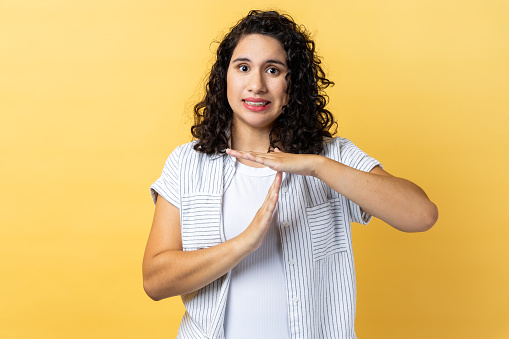 Portrait of bossy serious woman with dark wavy hair showing time out gesture, looking with strict expression at camera, deadline. Indoor studio shot isolated on yellow background.