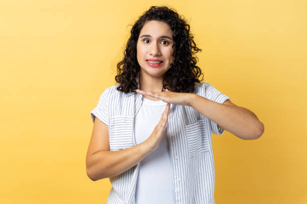 Woman showing time out gesture, looking with strict expression at camera, deadline. Portrait of bossy serious woman with dark wavy hair showing time out gesture, looking with strict expression at camera, deadline. Indoor studio shot isolated on yellow background. time out signal stock pictures, royalty-free photos & images