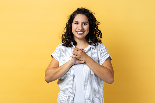 Portrait of positive optimistic friendly woman with dark wavy hair standing looking at camera, keeps hands together, expressing happiness. Indoor studio shot isolated on yellow background.