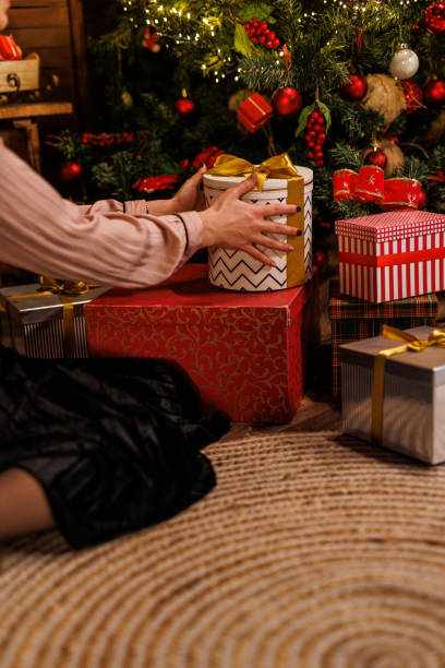 istock Young woman sitting on the floor, by the Christmas tree, stacking and arranging gift boxes for loved ones 1827393921