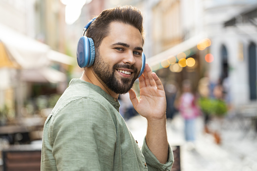 Rear view of man tourist in wireless headphones walking through the street outdoors, listening energetic music, dancing, turning to camera and smiling. Back view of guy traveler in urban sunshine city