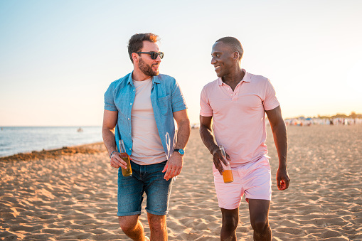 Smiling multi-ethnic males in their mid-adulthood, leisurely walking by the sandy shore, toasting with beer, and cherishing moments of fun during their seaside vacation.