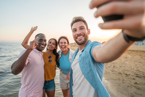 Mid adult individuals representing different ethnicities, capturing a joyful beach selfie with a smart phone.