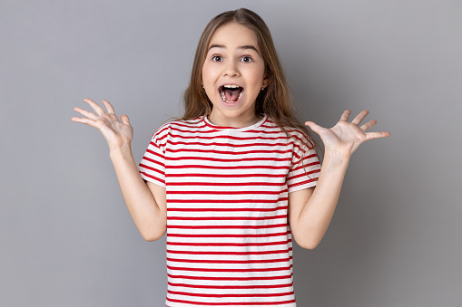 Portrait of excited little girl wearing striped T-shirt standing with raised arms and open mouth, hearing astonishing news, saying wow. Indoor studio shot isolated on gray background.