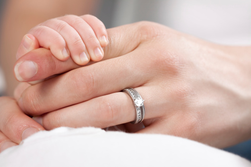 Small infant holding finger of mother who is wearing a diamond wedding ring.