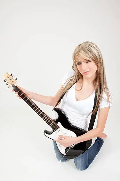 "A cute, blonde-haired, blue-eyed teenage girl poses in a studio, kneeling on the floor, a complex expression on her face. She looks directly into the camera, playing an electric guitar with its strap around her neck. Her right fingers rest on the upper frets of the guitar. Bare-foot, she wears a white, short-sleeved blouse and blue jeans. Her straight golden-blond hair cascades down her back. All against a light pink studio background. Her lips are pink."