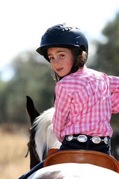 A young woman in western wear stands with her dark horse in a grassy pasture. Mountains and trees are in the background on a cloudy day in Europe.