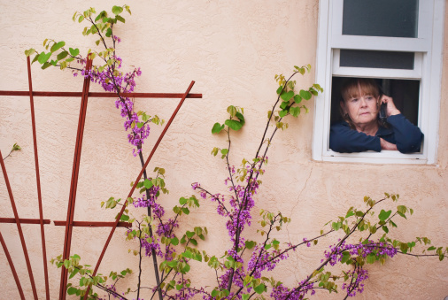 a senior woman looking pensive listening and talking on the telephone while leaning out the window of a residential house with purple plant and flower blooms.  horizontal composition.