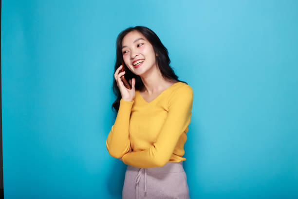 istock Asian woman poses while talking on the phone while standing in front of a light blue background. 1827516009
