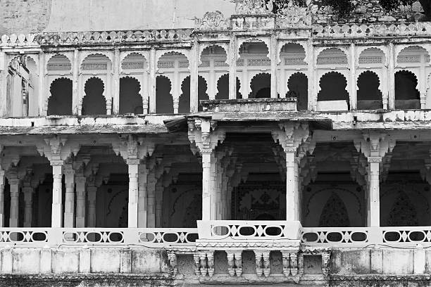 A straight-on, wide shot captures the iconic, elaborate facade of the Hawa Mahal palace. The upper stories showcase the characteristic terracotta-pink color, with a dense cluster of ornate jharokha (lattice) windows and small domes topped with golden finials. The base of the structure features ochre-colored walls and arched doorways, one containing a small stained glass window. The repetitive geometry and rich color define this historic architecture in Rajasthan.