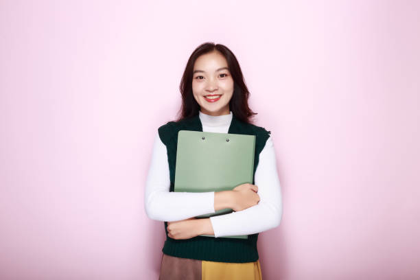 istock Asian woman poses holding a file folder while standing in front of a light pink background. 1827552898
