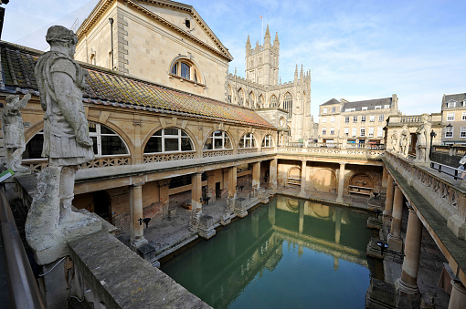 The Ancient Roman Baths in the English city of Bath - illuminated by afternoon sunshine casting reflections in the thermal bathBath Abbey is visible in the distance framed by the pillars and shadows of the ancient remains belowBritain