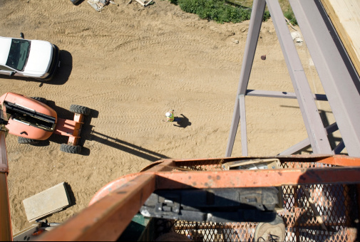 A construction worker walks across sand with track and tire marks on a job site along with a work truck and steel construction frame. Shot straight down from an aerial work platform 40aa above the ground. There are some sawdust shavings within the extreme right of the frame from the construction work being done on the platform.