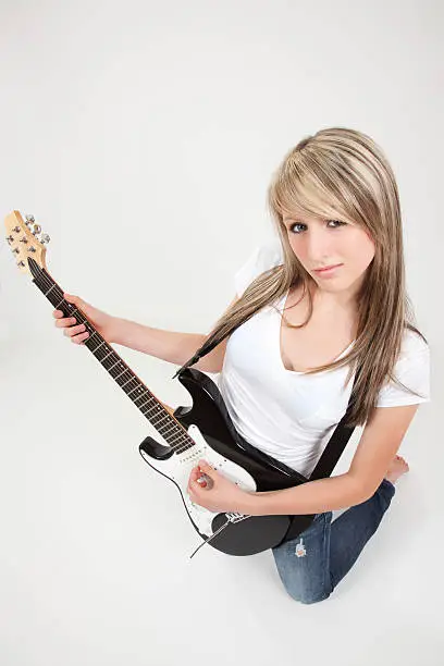 "A cute, blonde-haired, blue-eyed teenage girl poses in a studio, kneeling on the floor, only one leg showing, a complex expression on her face. She looks directly into the camera, playing an electric guitar with its strap around her neck. Her right fingers rest on the upper frets of the guitar. Bare-foot, she wears a white, short-sleeved blouse and blue jeans. Her straight golden-blond hair cascades down her back. All against a light pink studio background. Her lips are pink."