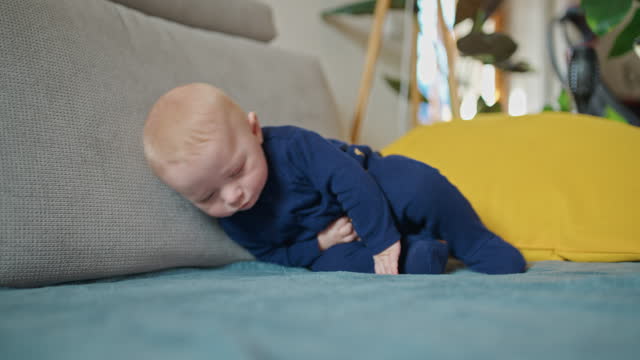 SLO MO Sleepy baby boy in blue velvet bodysuit falling upside down on sofa at home