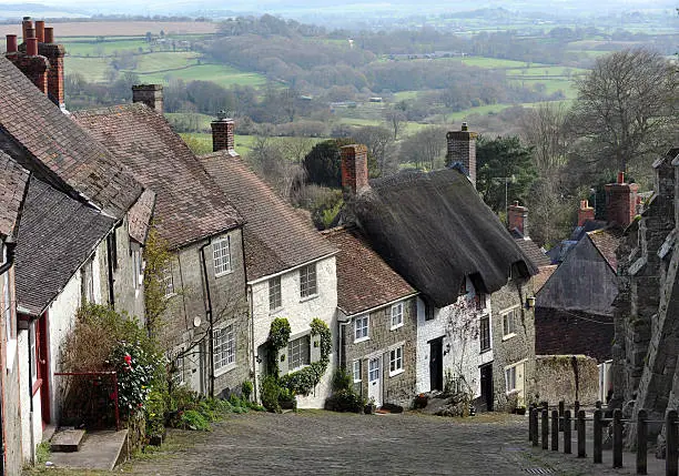 "An Iconic view of a row of quaint British Cottages built down a hill - and set against the rolling English countryside - in the West Country of EnglandGold Hill is one of the most generic images of England - made famous in an advert for Hovis BreadShaftesbury, Dorset, UK"