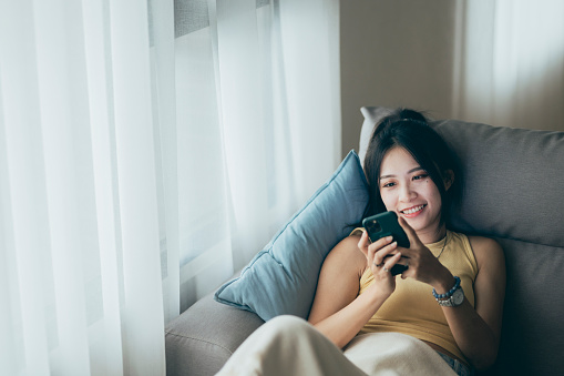 In a cozy living room, a young woman reclines on the sofa, absorbed in her smartphone. The soft glow of the screen illuminates her face as she engages with the device, her attention captured by the digital world at her fingertips.