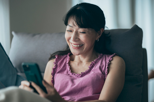 In the living room, a middle-aged woman lies comfortably on the sofa, engrossed in her smartphone. With leisurely swipes and taps, she navigates the Internet, possibly indulging in online shopping.