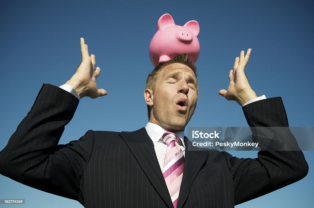 Businessman Tries to Balance Piggy Bank on Head Businessman tries to balance a pink piggy bank on his head in front of bright blue sky Financial Advisor Stock Photo