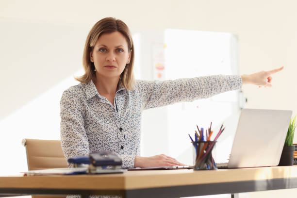 Angry businesswoman points finger to door asking to leave. Serious executive director sits at table near open laptop in office looking in camera