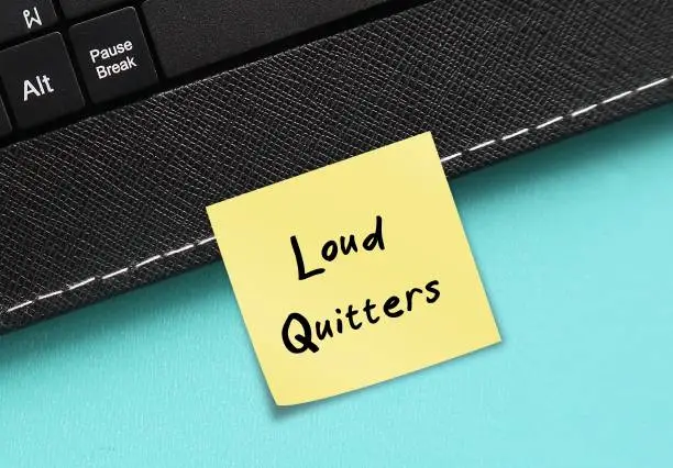 Photo of Keyboard on blue background with stick note written Loud Quitters - Unhappy workers employees who unlike quiet quitters, vocalizing their negative sentiments about organization before leaving