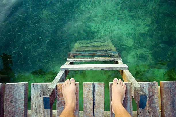 feet at the edge of a wooden dock with a ladder and clean waters below.