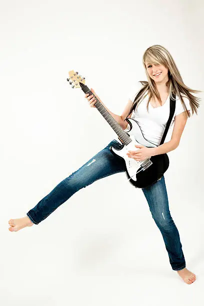 "A cute, blonde-haired, blue-eyed teenage girl poses in a studio. She looks directly into the camera, playing an electric guitar with its strap around her neck, while supported by only one leg. Her other leg is thrown outward, sideways, in a classic rock-'n-roll pose. Her right fingers rest on the upper frets of the guitar. Bare-foot, she wears a white, short-sleeved blouse and blue jeans. Her straight golden-blond hair cascades down her back. All against a light pink studio background. Her lips are pink."