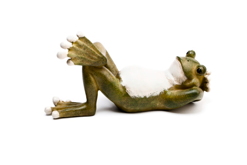 Green frog figurine relaxing on its back with its hands behind head. Studio shot on a white background.