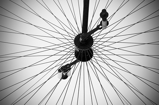 A close up shot of a still antique bike wheel, where the tire of the wheel cannot be seen. The wheel is made of many silver metal wires, and has a black and silver pedal. Behind the wheel is an empty grey background. The photos has a vignette effect. 