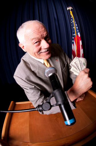 A corrupt politician smirks at a podium with a fistful of hundred-dollar bills.