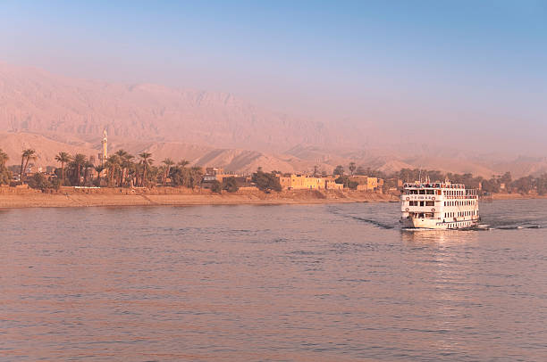 Nile Cruising Golden sunrise illuminates a typical Nile tourist cruise liner as it makes its way up the Nile in Egypt; Desert village backdrop nile river stock pictures, royalty-free photos & images