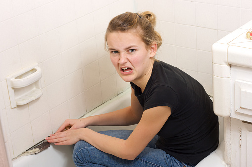 Young woman looks angry working to fix the bathtub caulk with toxic mold with a chisel and is involved in hard work and dirt