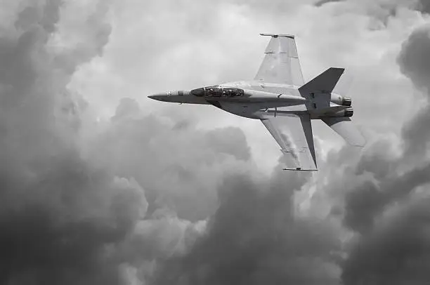 Desaturated photo of an F-18 Hornet cutting across a dramatic sky.