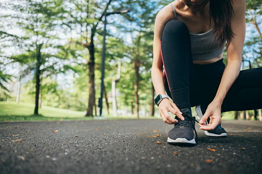 A young woman runner is outside in the morning, preparing for a jog. She is seen bending down, tying her shoelaces, ensuring her running shoes are securely fastened before she begins her run