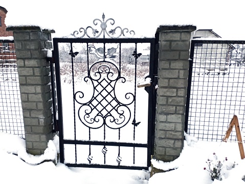 Metal gate in winter. Wrought metal fencing on the border between the yard and the street. Masonry columns made of concrete between which an entrance gate is installed.