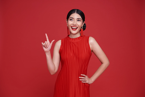 Happy Asian woman wearing red dress with hands pointing finger to copy space isolated on red background.